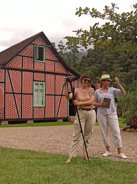 Fernanda do Canto, com câmera e tripé, e Carolina Bayer diante de uma casa enxaimel